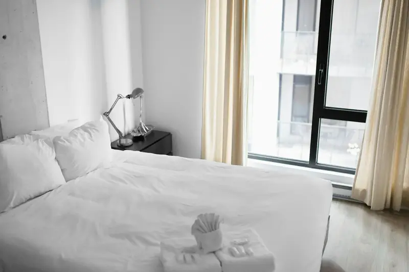 Made bed with white linens and folded towels next to a window